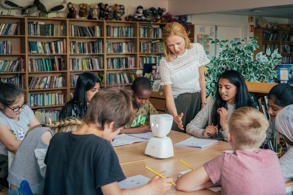 En kvinnelig lærer står og kommuniserer med åtte, ni unge elever som sitter rundt et bord. På bordet står roboten AV1.