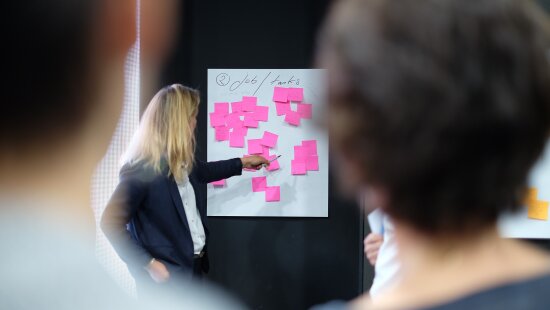 En dame står og peker mot en tavle med rosa post-it-lapper. To tilskuere med ryggen til fotografen ser mot tavlen.