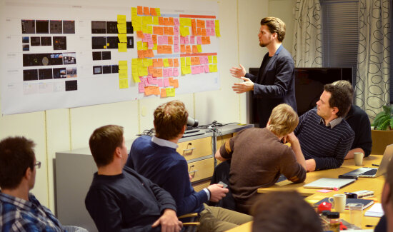 Workshop. Mennesker som sitter rundt et bord, mann ved tavla som snakker.