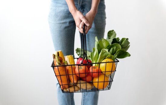Underkroppen på en kvinne kledd i lyseblå jeans. Hun holder en handlekurv full av frukt og grønnsaker i hendene.