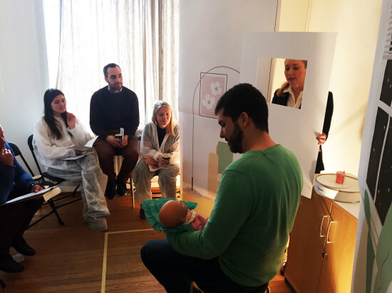 Flere mennesker sitter i et rom. En mann med grønn genser sitter med en babydukke i fanget. En dame står bak en skjerm av papir. Bildet illustrerer en designlab på et sykehus.