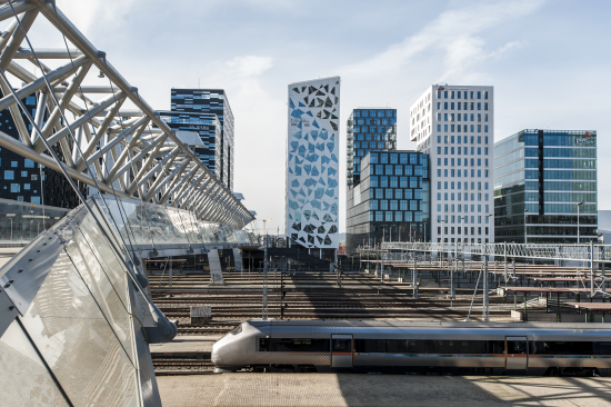 Flytoget som kjører ut fra Oslo sentralstasjon. Barcode-bygninger og Bjørvika i bakgrunnen.