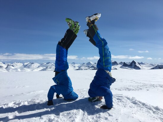 To menn i skiutstyr som står på hodet på toppen av et fjell. Blå himmel og snødekte fjelltopper i bakgrunnen.