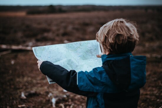 En liten gutt med ryggen til, blå vindjakke med hette. Han holder i et kart, og foran ham er et litt utvisket, jordfarget landskap.