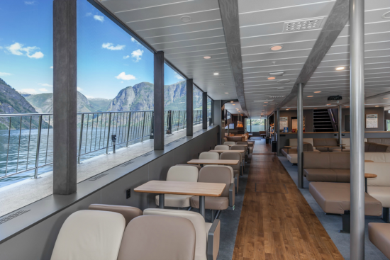 Interiøret i turistbåten Vision of the Fjords. Stoler og bord, parkettgulv, slanke søyler. Langs hele venstre side er det vinduer med utsikt til himmel med hvite skydotter, høye fjell og fjorden med småbølger.