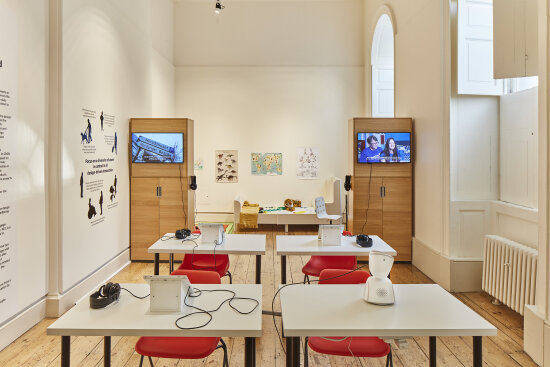 Et stort, luftig, hvitmalt rom, med høye buede vinduer. Rommet er innredet som et klasserom med fire pulter. Ved hver pult står en rød stol. På tre av pultene står det et nettbrett, og på den fjerde pulten står roboten AV1.