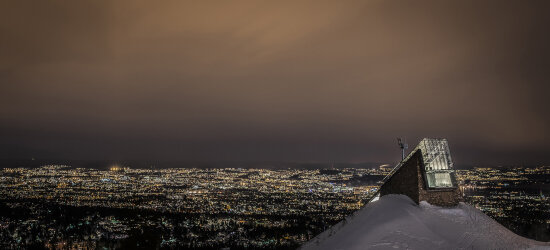 Hoppbakke (Midtstubakken, Holmenkollen nasjonalanlegg, Oslo. tegnet av ØKAW Arkitekter AS) om kvelden. Lys fra Oslo by i bakgrunnen.