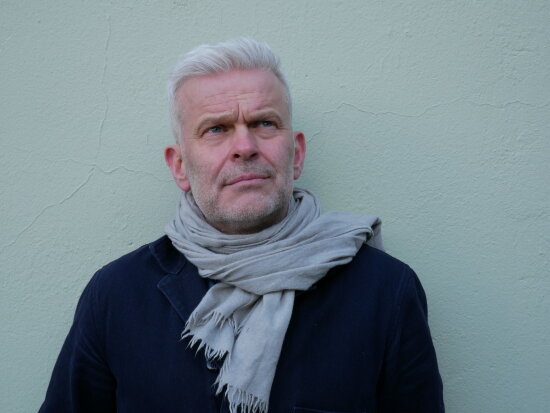 Portrett av Nils Stensrud mot en blågrønnn murvegg.