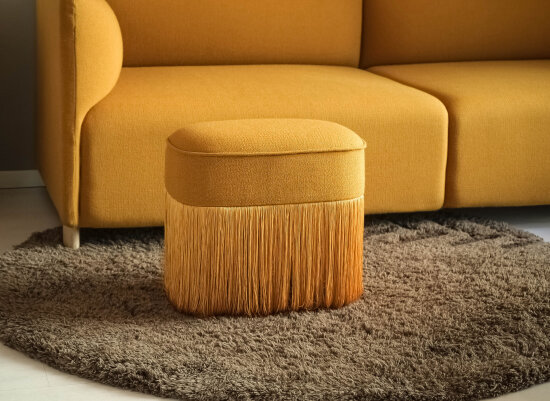 En gul, rund skammel/puff med lange frynser ned fra midten. En stor gul sofa i bakgrunnen.