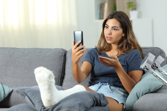 En jente sitter i en sofa. Hun har benet i gips og holder telefonen opp foran seg og snakker og gesikulerer mot den.