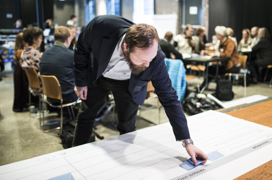 Mann med skjegg setter post it-lapp på stor plansje med mennesker sittende rundt bord i bakgrunnen.