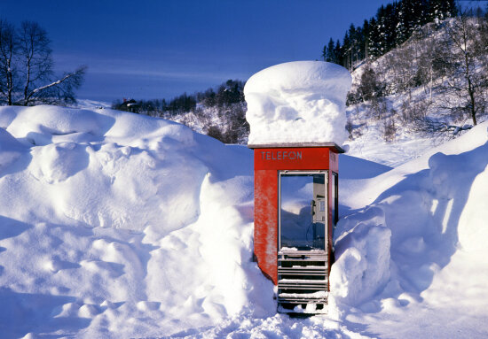 Rød telefonkiosk i meterdyp snø med et  tårn av snø på taket. I bakgrunnen blå himmel over et vinterlandskap av trær og snø.