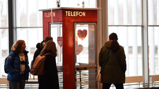 En rød telefonkiosk i et utstillingslokale. Innenfor vinduene henger hjerter hvor det står tallet 85. Fire ungdommer ser på telefonkiosken mens de går forbi.