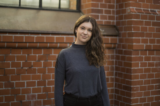 Portrett av Therese Stigen i halvfigur foran mursteinsvegg med vindu.