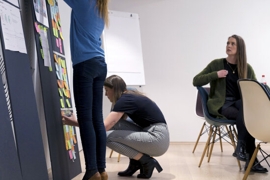 Tre kvinner i workshop. To klistrer post-it-lapper på tavle, en sitter ved bord og følger med.